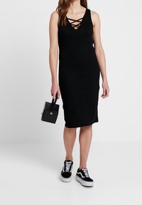 Schwarzes ärmelloses Kleid mit tiefem V-Ausschnitt und überkreuzten Trägern. Kombiniert mit schwarzen Sneakern und einer kleinen schwarzen Handtasche.