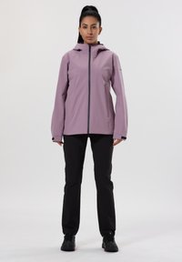 Giacca viola antivento con cappuccio, zip frontale e due tasche. Abbinata a pantaloni neri e calzature nere, su uno sfondo neutro.