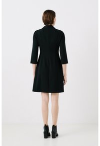 Robe noire ajustée avec des manches trois-quarts, un col et une fermeture éclair au dos. Le tissu semble lisse avec une silhouette structurée.