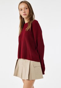Burgundy neulepaita, jossa on väljä malli ja pyöreä pääntie, yhdistettynä vaaleanbeigeen laskostettuun hameeseen, jossa on suuret sivutaskut.