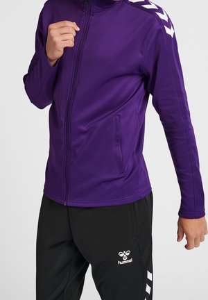 Giacca viola con cerniera e texture liscia, con accenti bianchi a chevron sulle spalle e sulle tasche laterali. Abbinate a pantaloni neri.