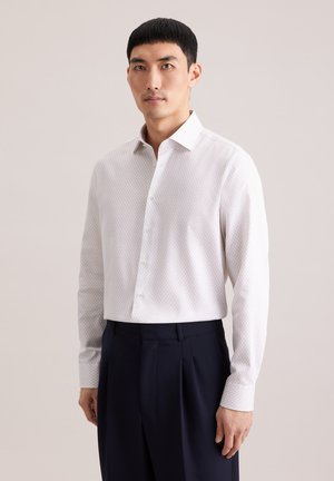 Uomo che indossa una camicia bianca a maniche lunghe con motivi, infilata in pantaloni neri plissettati, in piedi davanti a uno sfondo chiaro.