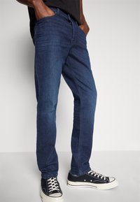 Jeans din denim albastru închis, cu un design drept, trei buzunare laterale și o ușoară decolorare. Asortați cu teniși negri din pânză.