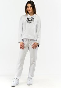 Sudadera blanca con estampado "ICE", de corte amplio y con cordones, combinada con pantalones ajustados a juego; ambos hechos de un tejido suave. Zapatillas blancas completan el look.