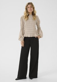 Donna con lunghi capelli biondi ondulati, che indossa un top beige a maniche lunghe con arricciature e pantaloni neri a gamba larga, in piedi con una mano in tasca.