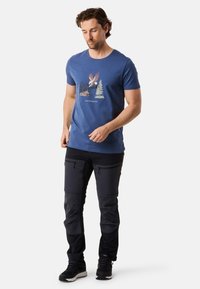 Blå bomullst-shirt med ett grafiskt tryck av en örn och träd. Svarta cargo-byxor med dragkedjor. Svarta sneakers med vita sulor.