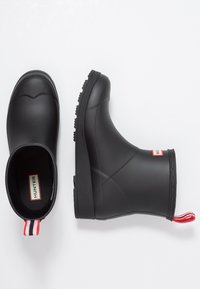 Bottes de cheville en caoutchouc noir avec des semelles texturées, bout rond et une étiquette rouge et blanche à l'arrière. L'intérieur présente une étiquette de marque.