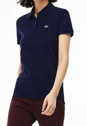 Femme portant un polo bleu marine ajusté à manches courtes avec un petit logo crocodile vert sur la poitrine et un pantalon foncé.
