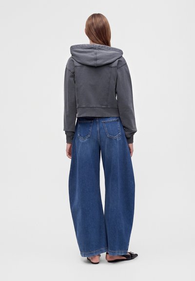 Femme aux longs cheveux portant un sweat à capuche gris foncé et un jean bleu à jambes larges, debout de dos sur un fond clair uni.