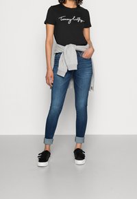 Camiseta negra con el logo blanco de "Tommy Hilfiger", suéter gris claro anudado alrededor de la cintura, jeans ajustados azules y zapatillas negras sin cordones.