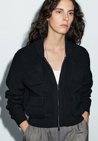 Woman with long curly dark hair wearing a black knitted zip-up cardigan over a white top and gray plaid pants, hands in pockets.