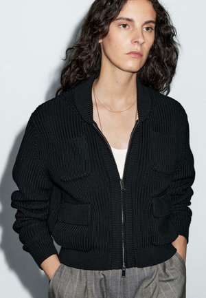 Frau mit langen lockigen dunklen Haaren, die eine schwarze gestrickte Zip-Up-Strickjacke über einem weißen Oberteil und grauen karierter Hose trägt, mit den Händen in den Taschen.