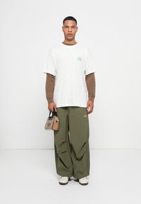 Witte oversized t-shirt met groen logo, bruin lang shirt eronder, olijfgroene wijde broek en kleine beige tas met zwart adidas logo.
