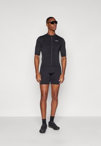 Combinaison de cyclisme noire à manches courtes et à fermeture éclair. Le matériau est lisse et ajusté, associé à des chaussures de cyclisme noires et des lunettes de soleil.