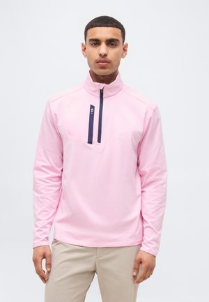 Giovane uomo che indossa un maglione a maniche lunghe rosa chiaro con una cerniera verticale nera sul petto e pantaloni beige, in piedi su sfondo bianco.