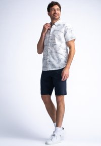 Camisa de botones de manga corta en tela ligera con fondo blanco y estampado de inspiración natural en gris, combinada con pantalones cortos azul marino y zapatos blancos.