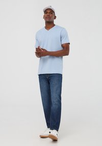 Maglietta azzurra chiara a V, maniche corte, abbinata a jeans blu scuro e sneakers bianche con dettagli neri. Design semplice, vestibilità casual.