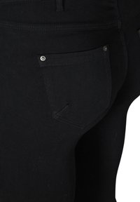 Jean en denim noir avec une vue en gros plan de la poche arrière. Les caractéristiques comprennent un clou argenté près de la poche, une texture lisse et un design ajusté.