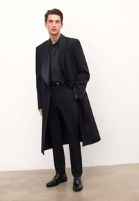 Abrigo negro con solapas de satén, usado sobre una camisa oscura y pantalones a medida. Zapatos de vestir negros pulidos. Textura suave, diseño estructurado.