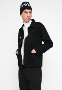 Mustast teksariidest jakk ees taskute, lukuga trukkidega ja veidi lõdva lõikega, kombineeritud valge kõrge kaelusega džempri ja musta mütsiga.