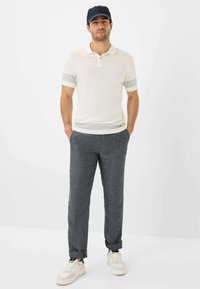 Polo bianco con strisce grigie, abbinato a pantaloni di lino grigio scuro arrotolati. Modello che indossa un cappellino blu navy e sneakers bianche.