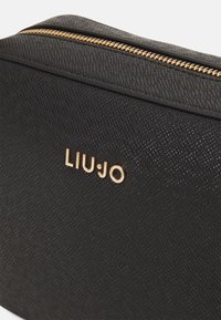 Geantă sintetică texturată neagră, cu un fermoar aurit și numele brandului „LIU•JO” embosat pe față.
