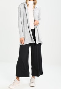 Grå hoodiekofta med långa ärmar, mjuk textur, öppen frontdesign, kombinerad med svarta vida byxor och ljusa sneakers.