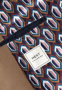 Tela de seda con un patrón geométrico en tonos azules, rosas y marrones. Presenta una etiqueta cosida con "NEXT TAILORING".