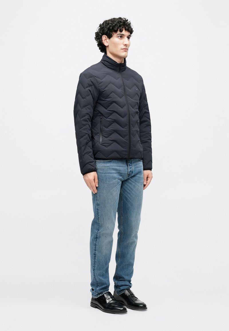 Veste matelassée noire avec col montant, présentant un motif diagonal et des poches zippées, associée à un jean bleu et des chaussures noires.