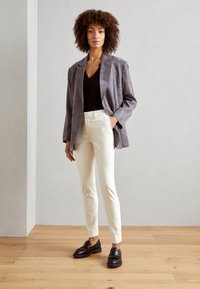 Blazer en daim gris porté sur un haut noir, associé à un pantalon blanc sur mesure et des mocassins sombres, le tout sur un fond clair avec un sol en bois.