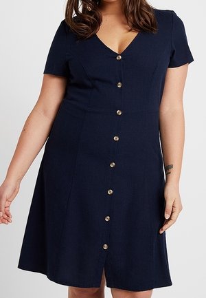 Mujer vistiendo un vestido azul marino de manga corta con escote en V y cierre frontal de botones, posando frente a un fondo liso.