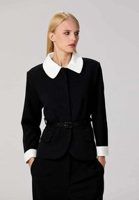 Veste noire sur mesure avec col et poignets blancs, taille ceinturée, deux poches avant et une silhouette structurée. Texture lisse.