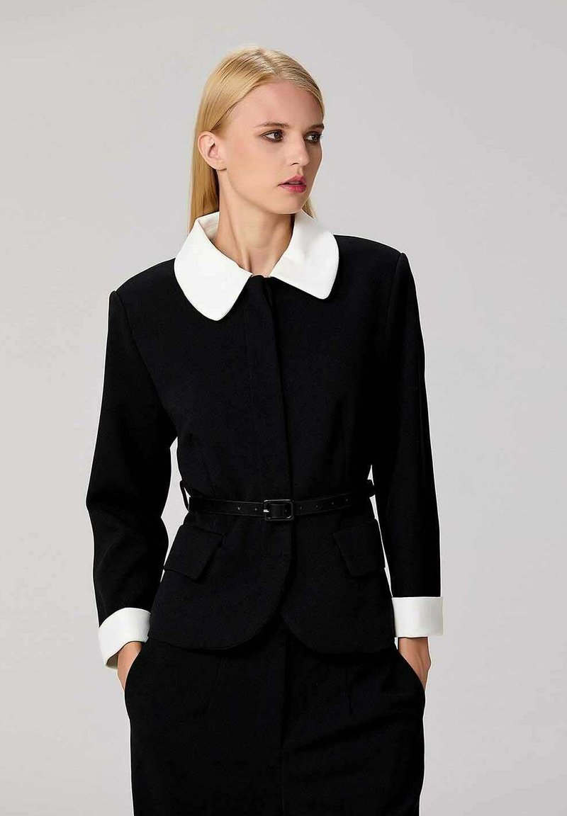 Veste noire sur mesure avec col et poignets blancs, taille ceinturée, deux poches avant et une silhouette structurée. Texture lisse.