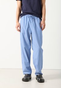 Vyras, vilkintis laisvus mėlynai baltais dryžuotus raištelių kelnes, juodus slip-on batus ir tamsiai mėlyną marškinėlį, stovintis ant šviesios grindų.