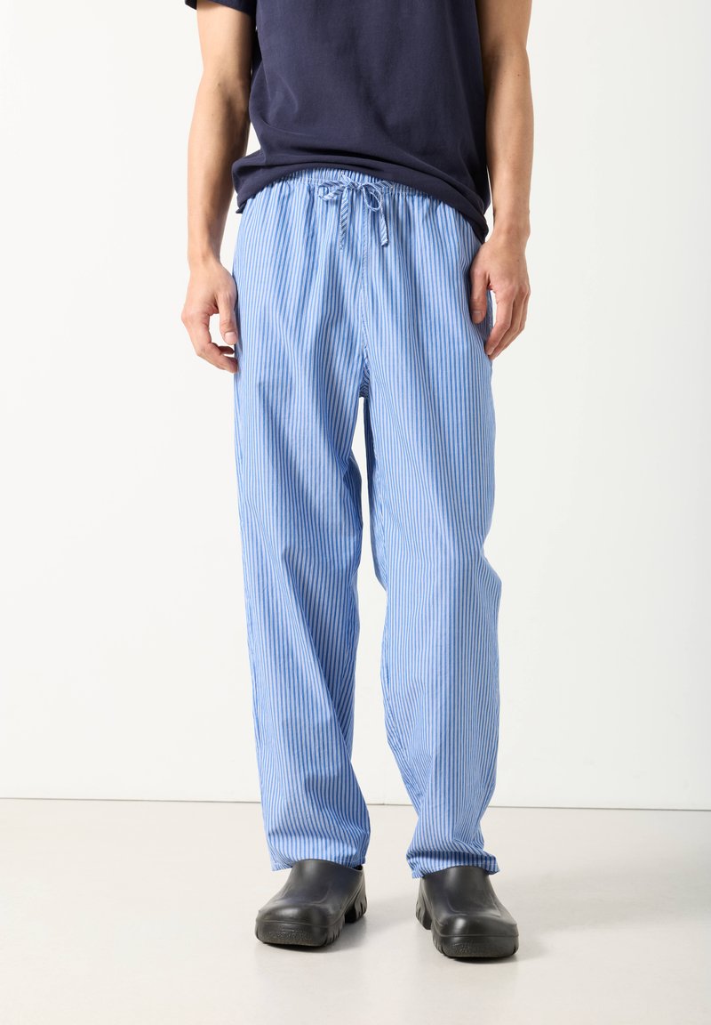 Vyras, vilkintis laisvus mėlynai baltais dryžuotus raištelių kelnes, juodus slip-on batus ir tamsiai mėlyną marškinėlį, stovintis ant šviesios grindų.