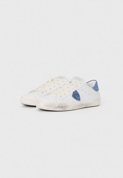 Białe sneakersy z jasnoniebieskimi akcentami, teksturowana tkanina i brązowe sznurowadła. Posiadają niebieską łatę po boku oraz zużytą białą podeszwę.