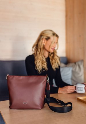 Hnědá syntetická kožená kabelka přes rameno se stříbrným kováním, odnímatelným popruhem a detaily loga na přední straně.