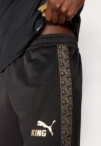 Musta urheiluhousut, joissa on kultaiset yksityiskohdat, logo ja sana "KING". Housuissa on kuvioitu sivupaneeli ja joustava vyötärönauha.