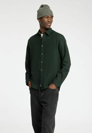 Camisa abotonada de color verde oscuro con tela texturizada, cuello redondo y mangas largas, combinada con jeans negros y un gorro de lana gris.