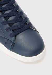 Navyblaue Sneakers mit strukturiertem synthetischen Leder, dicken passenden Schnürsenkeln und weißer Gummisohle vor einem weißen Hintergrund.