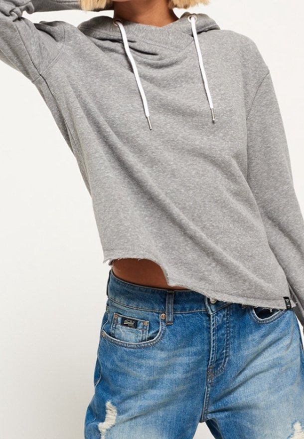 Sudadera gris recortada con capucha con cordón, que presenta mangas largas y un dobladillo sin rematar. Combinada con unos jeans de mezclilla azul desgastados.