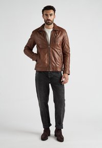 Bruine leren jas met gewatteerde schouderaccenten, rits aan de voorkant, twee zijzakken, gedragen met een donkere spijkerbroek en bruine suède laarzen.