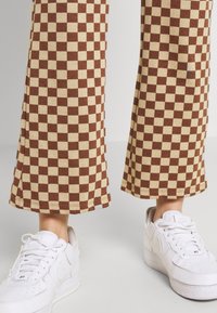 Bruna och cremefärgade rutiga byxor med lös passform och utsvängd nederdel, kombinerade med vita sneakers. Tyget verkar mjukt och texturerat.