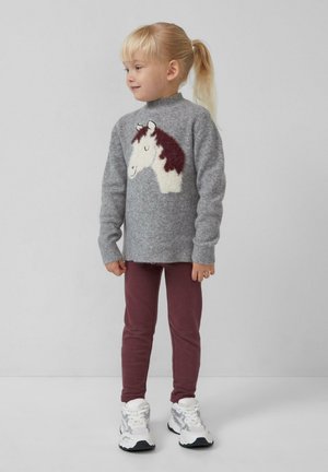Junges Kind mit blonden Haaren, das zu einem Ponytail gebunden ist, trägt einen grauen Pullover mit einem Pferdegrafik, maroonfarbene Hosen und weiße Turnschuhe und steht vor einem schlichten Hintergrund.
