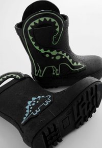 Bottes en caoutchouc noires ornées de motifs de dinosaures verts, surface texturée et semelle à motif. Des gouttes d'eau sont visibles sur la surface.