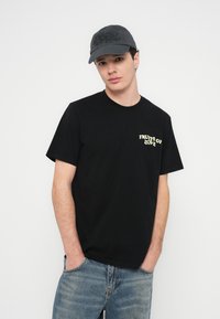 T-shirt preta de algodão com mangas curtas e gola redonda, com a frase "FRUITS OF LOVE" impressa a amarelo no lado esquerdo do peito. Calças de ganga azul claro.