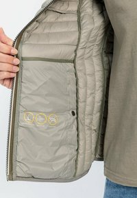 Zielona, ocieplana kurtka od wewnątrz z pikowaną strukturą. Posiada ikony odporności na wodę i wiatr. Zbliżenie na detale zapięcia na zamek i guziki.