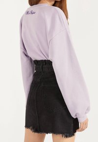 Fialový oversized svetr s kleslými rameny a fialovým textem na zadní straně, spárovaný s černou denimovou mini sukní s třepeným lemem.