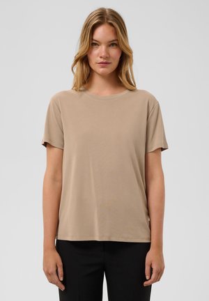 Frau mit blondem Haar, die ein beiges Kurzarmshirt und schwarze Hose trägt, steht vor einem schlichten hellen Hintergrund.