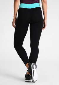 Czarn Legginsy z turkusowym pasem, wykonane z elastycznego materiału. Posiadają dopasowany krój oraz czarne buty sportowe z białą podeszwą.